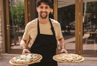 Man met zwart schort en beige shirt houdt twee pizza’s bij Wecamp Cala Montgó vakantiepark in Catalonië.