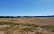 Blick auf eine trockene Wiese mit Sträuchern bei einer Lodge, blauer Himmel und See im Hintergrund.