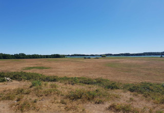 Foto van een droog veld met verspreide struiken bij een lodge, blauwe lucht en een meer in de verte.