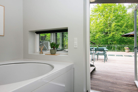 Badezimmer mit Badewanne und Blick auf eine Terrasse mit Gartenmöbeln und Bäumen in Ebeltoft, Dänemark.