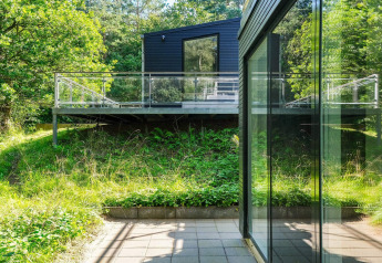 Moderne sort-hytte med stor terrasse og glasrækværk omgivet af frodig natur i Ebeltoft, Danmark.