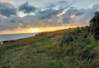Coucher de soleil près d’un lodge avec herbe, rivage rocheux et grands nuages éclairés par la lumière dorée.