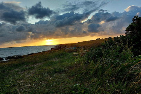 Avondzon bij een lodge, met grasland en stenen aan de kust, onder een lucht vol dramatische wolken.
