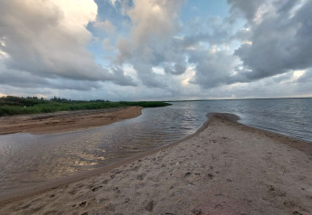 Udsigt fra en lodge over en sandstrand med vandløb, siv og dramatiske skyer i baggrunden.