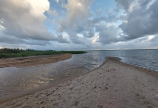 Vue depuis un lodge sur une plage de sable, de l’eau, des roseaux et un ciel nuageux spectaculaire.