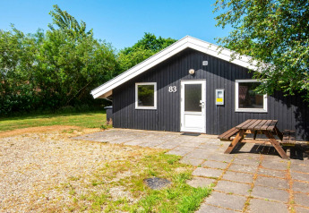 Hyggelig sort træhytte ved navn Secluded Harbor Cottage med udendørs bænk, omgivet af grønt i Danmark.