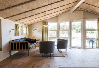 Salon lumineux avec plafond en bois incliné, grandes fenêtres et mobilier moderne à Blåvand, Danemark.
