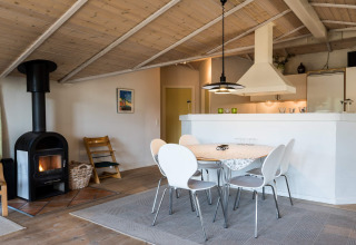 Zona soggiorno accogliente con tavolo da pranzo, stufa a legna e cucina aperta in casa vacanze a Blåvand, Danimarca.