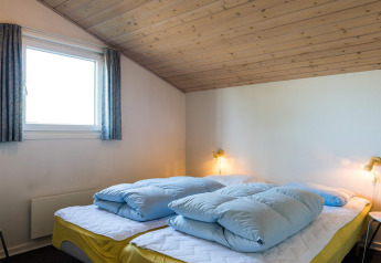 Soveværelse med skråt træloft, to senge med gule lagner og blå dyner på feriebolig i Blåvand, Danmark.