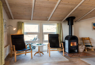 Wohnbereich im Ferienhaus in Blåvand, Dänemark, mit Kaminofen, Holzdecke, Sesseln und Fenster.