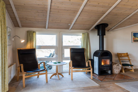 Woonkamer in vakantiehuis in Blåvand, Denemarken, met houtkachel, stoelen en houten plafond.