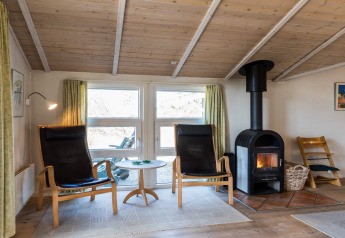 Stue i feriebolig i Blåvand, Danmark, med træloft, brændeovn, to sorte stole og store vinduer.