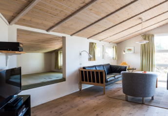 Stue i feriebolig i Blåvand, Danmark med træloft, sofa, lænestole og stort vindue med grønne gardiner.