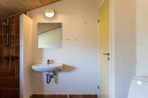 Badezimmer in einem Ferienhaus in Blåvand mit Waschbecken, Spiegel, Dusche und schräger Holzdecke.