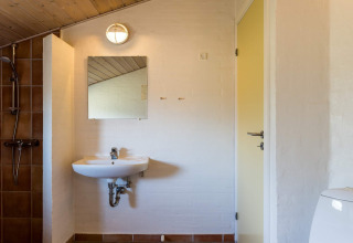 Badezimmer in einem Ferienhaus in Blåvand mit Waschbecken, Spiegel, Dusche und schräger Holzdecke.