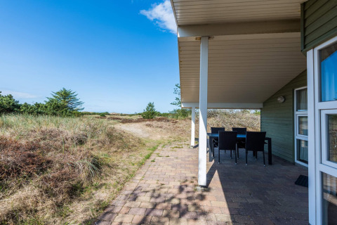 Patio coperto con tavolo da pranzo in una casa vacanze a Blåvand, Danimarca, circondata da dune e verde.
