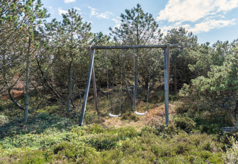 Gyngestativ med to gynger placeret i en grøn skov nær feriehytte i Blåvand, Danmark, under blå himmel.