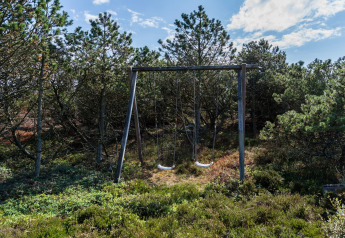 Gyngestativ med to gynger placeret i en grøn skov nær feriehytte i Blåvand, Danmark, under blå himmel.