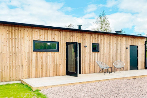 Moderne svensk lodge med træfacade, sort dør, to stole og bord på træterrasse under blå himmel.