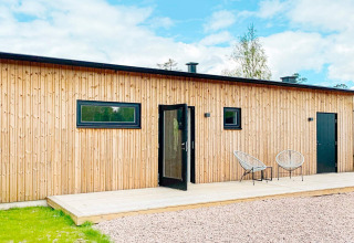 Moderne svensk lodge med træfacade, sort dør, to stole og bord på træterrasse under blå himmel.