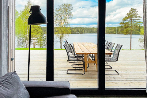Udsigt fra en stue med sofa og lampe mod en træterrasse med bord og stole ved en sø i Sverige.