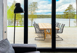Udsigt fra en stue med sofa og lampe mod en træterrasse med bord og stole ved en sø i Sverige.