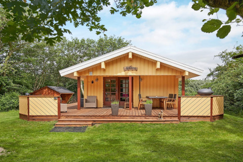 Hyggelig kysthytte-retreat i Danmark med træterrasse og grøn græsplæne omgivet af frodige træer.