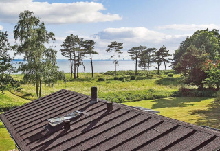 Vue d'une maison de vacances à Knebel, Danemark, avec toit, herbe, arbres et mer à l'arrière-plan.