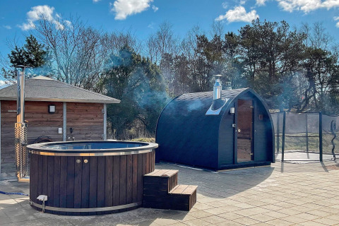 Udendørs spa og sauna ved feriehus i Sæby, Danmark, omgivet af træer og blå himmel.