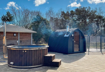Buitenspa en sauna bij een vakantiehuis in Sæby, Denemarken, met bomen en blauwe lucht.