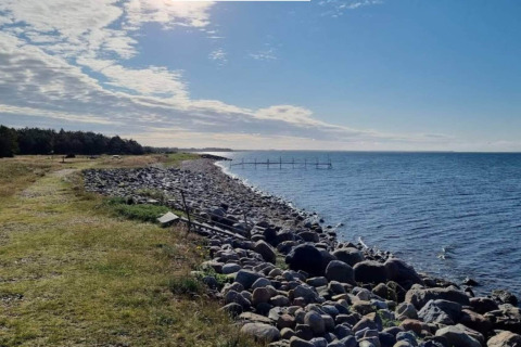 Kystudsigt fra Cozy Coastal Cottage Retreat i Danmark, med stenstrand, blåt hav og grønt landskab.