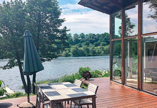 Wooden deck with dining table, chairs and umbrella overlooking a lake near a glass-walled lodge in Bryrup, Denmark.