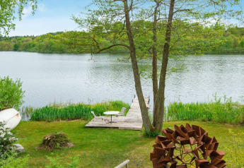 Lakeside wooden dock with table and chairs, surrounded by greenery at a holiday home in Bryrup, Denmark.