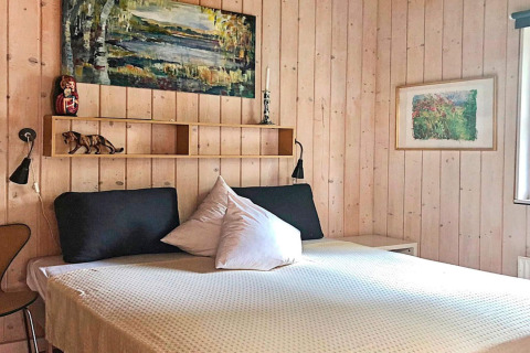 Schlafzimmer im Ferienhaus in Bryrup, Dänemark, mit Holzwänden, gemütlichem Bett, Bildern und Regal.