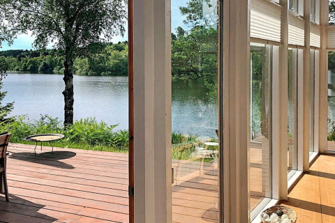 View from lodge in Bryrup, Denmark, with wooden deck, large windows, and scenic lake and trees.
