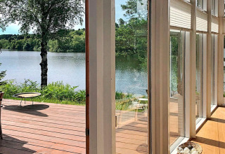 View from lodge in Bryrup, Denmark, with wooden deck, large windows, and scenic lake and trees.