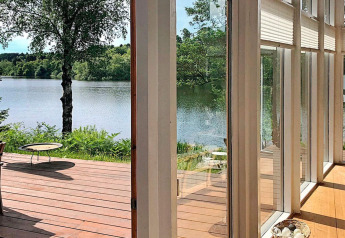 Zicht uit lodge in Bryrup, Denemarken, met houten terras, grote ramen en een mooi meer.