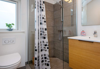 Baño moderno con ducha, inodoro, ventana y lavabo de madera en casa de vacaciones para 8 personas en Sydals, Dinamarca.