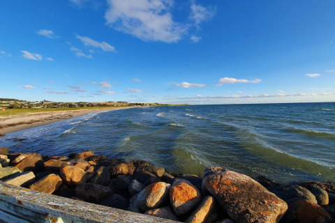 Udsigt over stranden og havet fra en hytte i Knebel, Danmark, med sten i forgrunden og blå himmel.