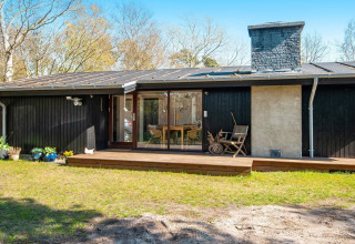 Sort træhytte med terrasse og have, set udefra på en solskinsdag i Knebel, Danmark, med moderne detaljer.