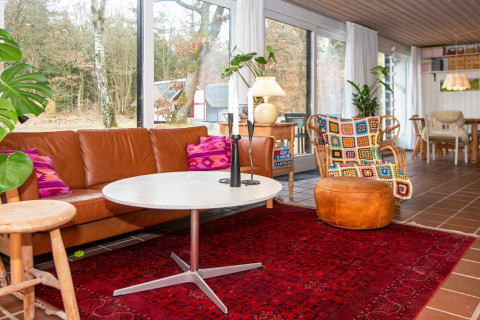 Salon d'une maison de vacances à Ebeltoft, Danemark, avec canapé en cuir, coussins colorés et grandes fenêtres.