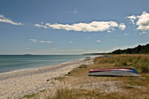Peaceful sandy beach with overturned boats, grassy shore, calm sea and a partly cloudy sky in sunlight.