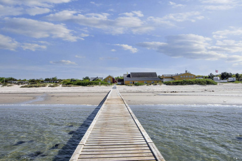 Houten pad naar geel vakantiehuis op het strand in Glesborg, Denemarken, onder blauwe hemel.