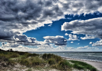 Ciel dramatique et nuageux sur la plage près d’un lodge, avec herbes, mer agitée et lumière contrastée.