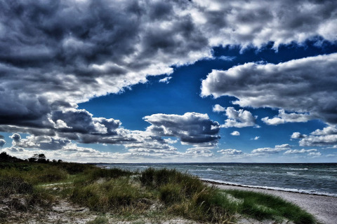 Dramatic sky with dark clouds over a beach by a lodge, featuring grass, sand, and waves in the distance.