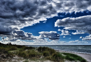 Dramatic sky with dark clouds over a beach by a lodge, featuring grass, sand, and waves in the distance.