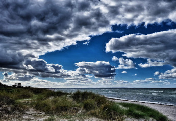 Cielo dramático con nubes oscuras sobre una playa junto a una cabaña, con hierba, arena y olas de fondo.