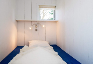 Narrow bedroom with single bed, blue mattress, white pillows, wall lamps and window at Glesborg lodge.