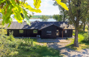 Vakantiehuis in Millinge, Denemarken, met bomen rondom en een meer op de achtergrond, zonnige sfeer.
