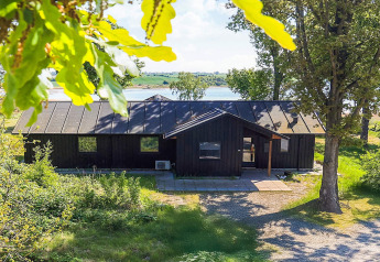 Vakantiehuis in Millinge, Denemarken, omgeven door bomen, meer op de achtergrond, zonnige sfeer.